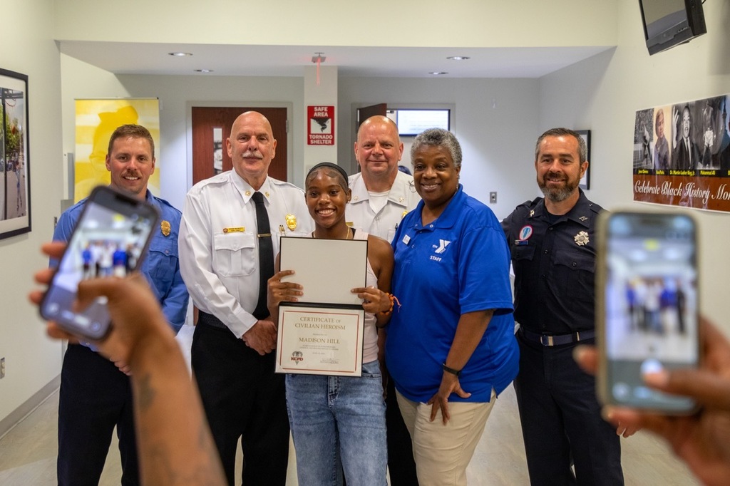 Madison Hill holds her civilian heroism award from KCFD, surrounded by members of the department as she smiles for a picture
