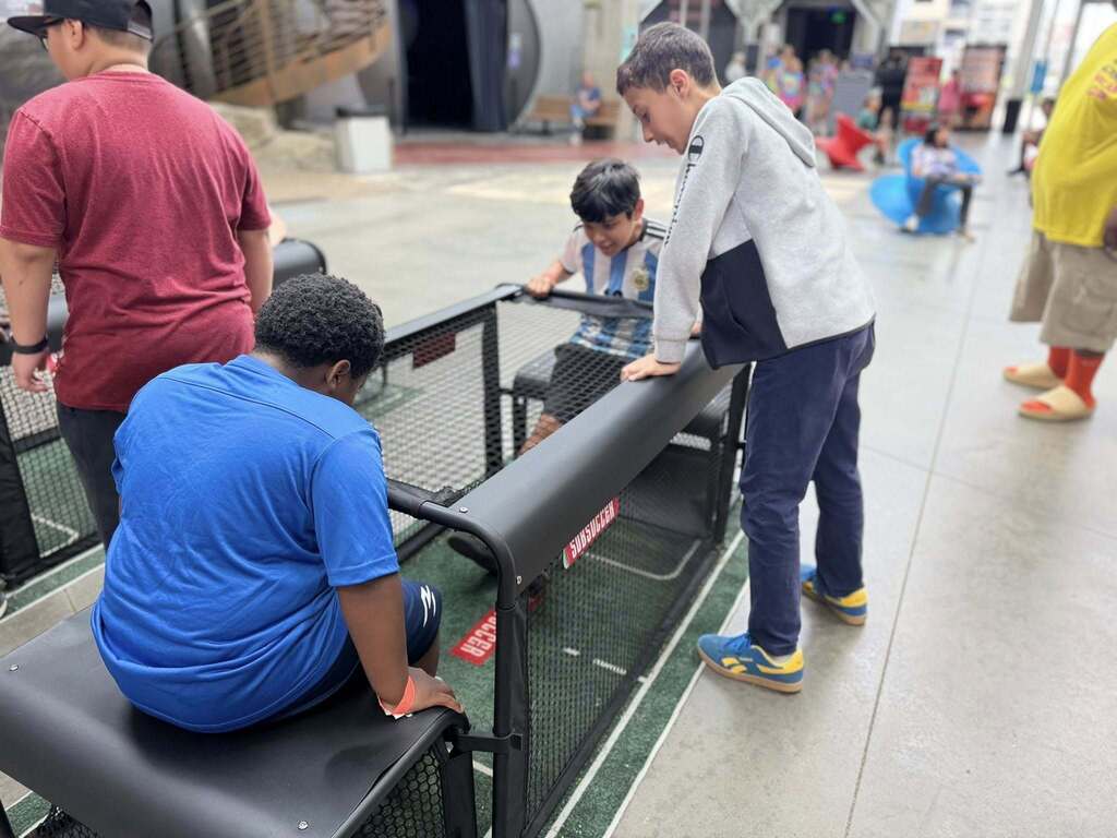 Two students sit at an interactive display while another student stands next to them watching