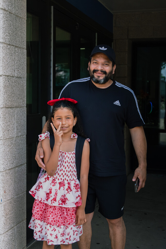 A man in a black t-shirt, shorts, and hat stands smiling at the camera with his arm wrapped around his young daughter who wears a red and white patterned dress is smiling, holding a peace sign in front of her face
