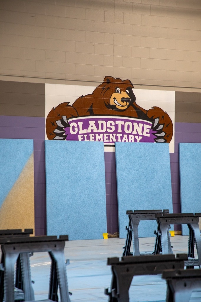 Saw horses are visible in the foreground with pieces of sound-minimizing material in the background propped up against a wall. On the wall, a mural reads "gladstone elementary" with the school's grizzly bear mascot behind it.