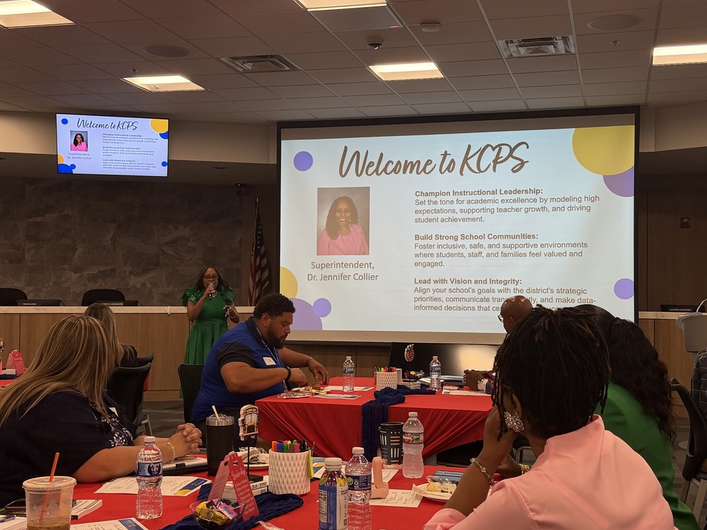 A woman stands in front of a screen projector with a slide that says "welcome to kcps" and in the foreground people are seated at tables with plates of food in front of them