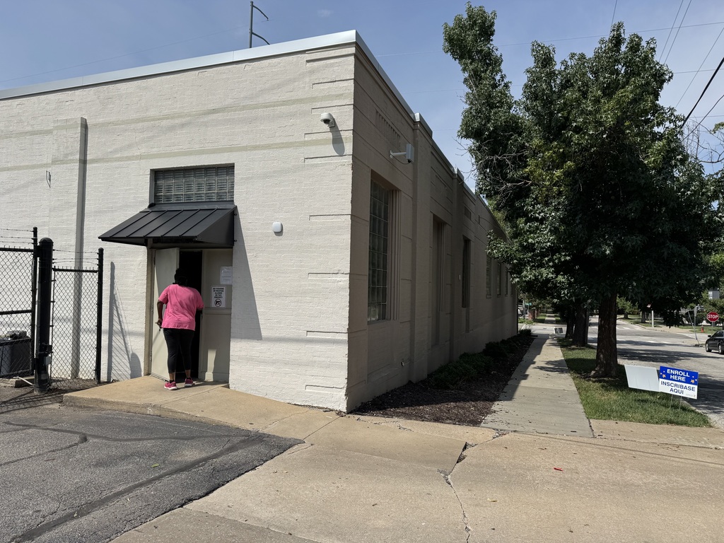 A woman in a bright pink t-shirt walks into a building shaded by trees on the side facing the street