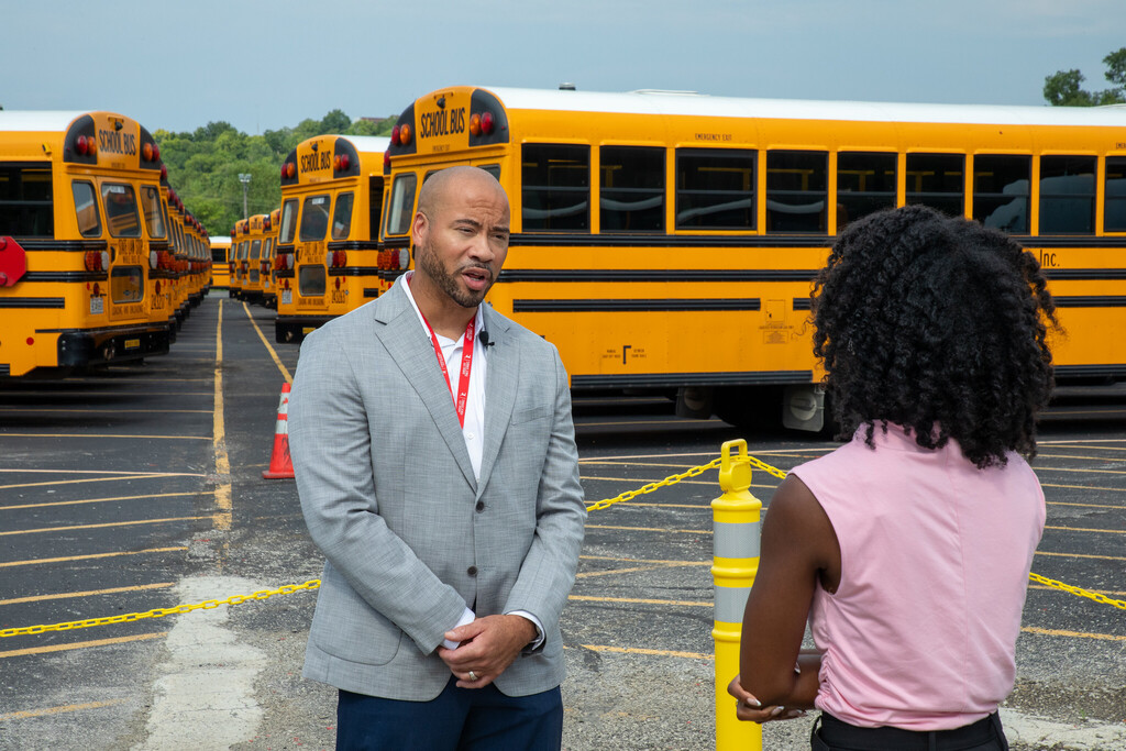 A man facing toward the camera speaks to a female reporter who has her back to the camera as she interviews him. School buses are visible in the background