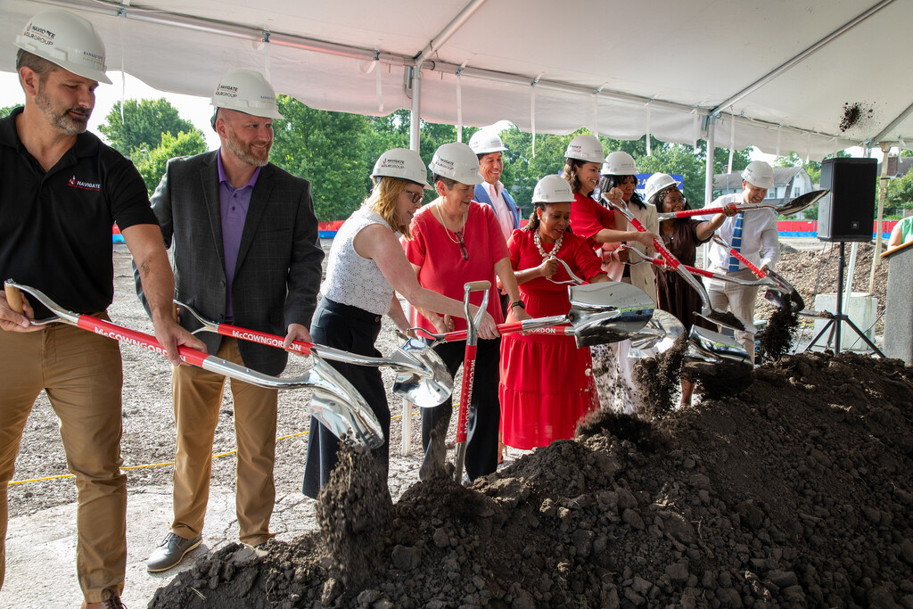 A line of adults wearing white construction helmets use ceremonial shovels to scoop dirt at a groundbreaking ceremony