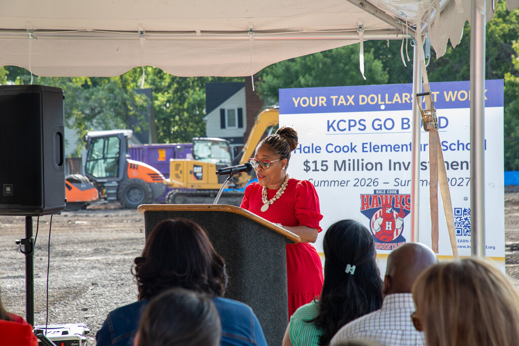 superintendent dr. jennifer stands at a podium with her hair up in a bun and wearing a red dress. She is looking down at her notes as she speaks to a crowd in front of her