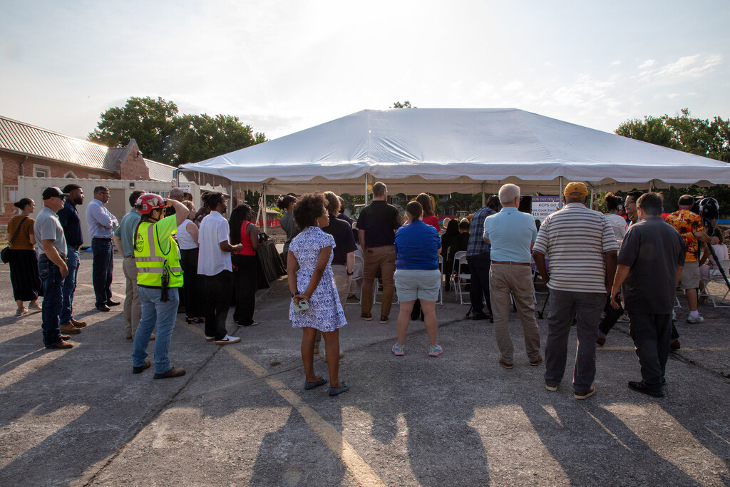 a moderate crowd of adults stand with their backs to the camera as they watch the proceedings of a groundbreaking ceremony happening in front of them