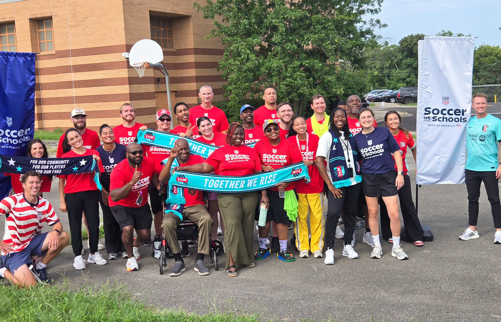 A group of adults wearing red t-shirts gather and smile at the camera. Some hold teal scarves that say "together we rise" with the KC Current team logo on either side