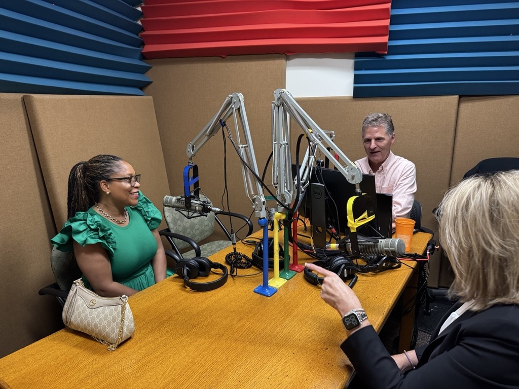 A woman in a green dress, a man in a light pink dress shirt, and a woman in a black blazer sit around a table, each with a tall radio microphone positioned in front of them