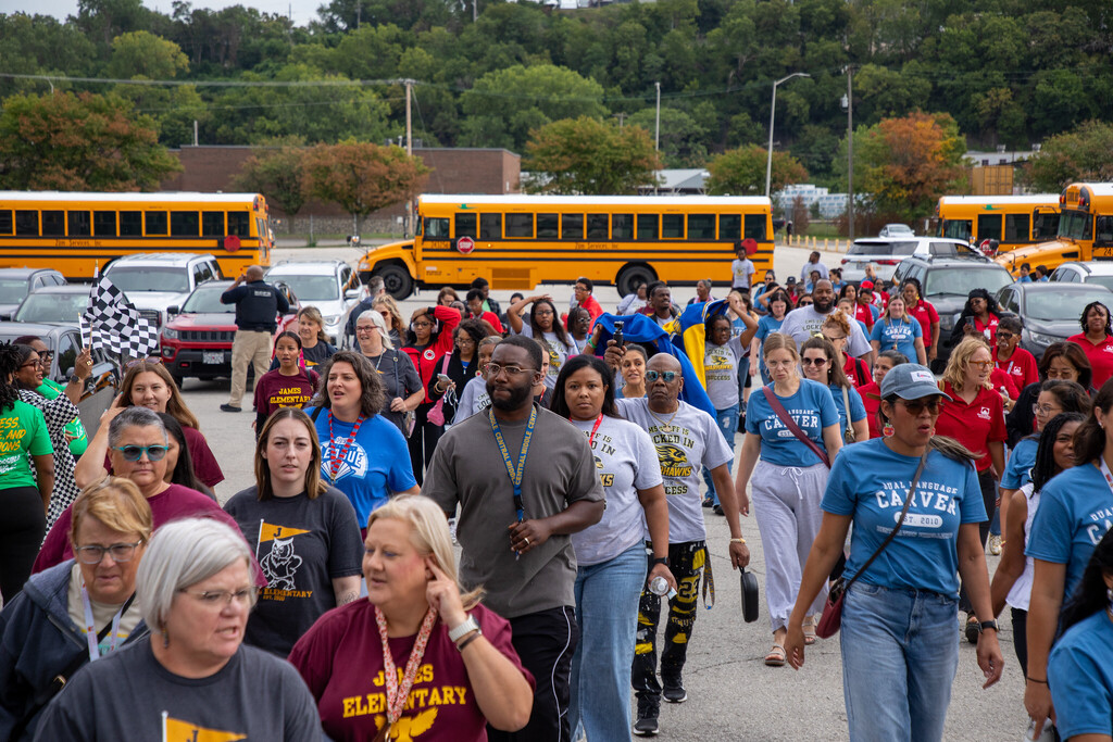 A large crowd of adults walks toward the camera with school buses visible in the distant background
