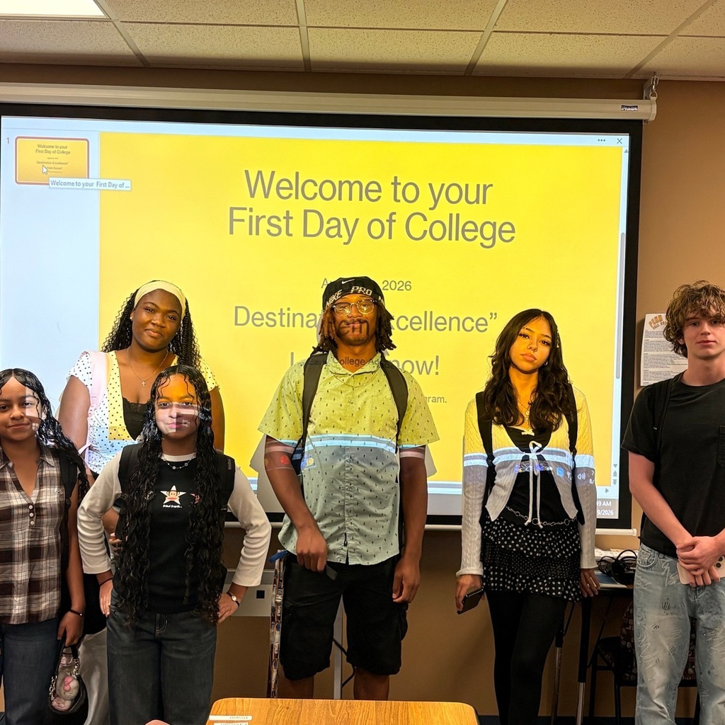 Six students stand in a line looking at the camera in front of a projector screen that has "welcome to your first day of college" projected on to it