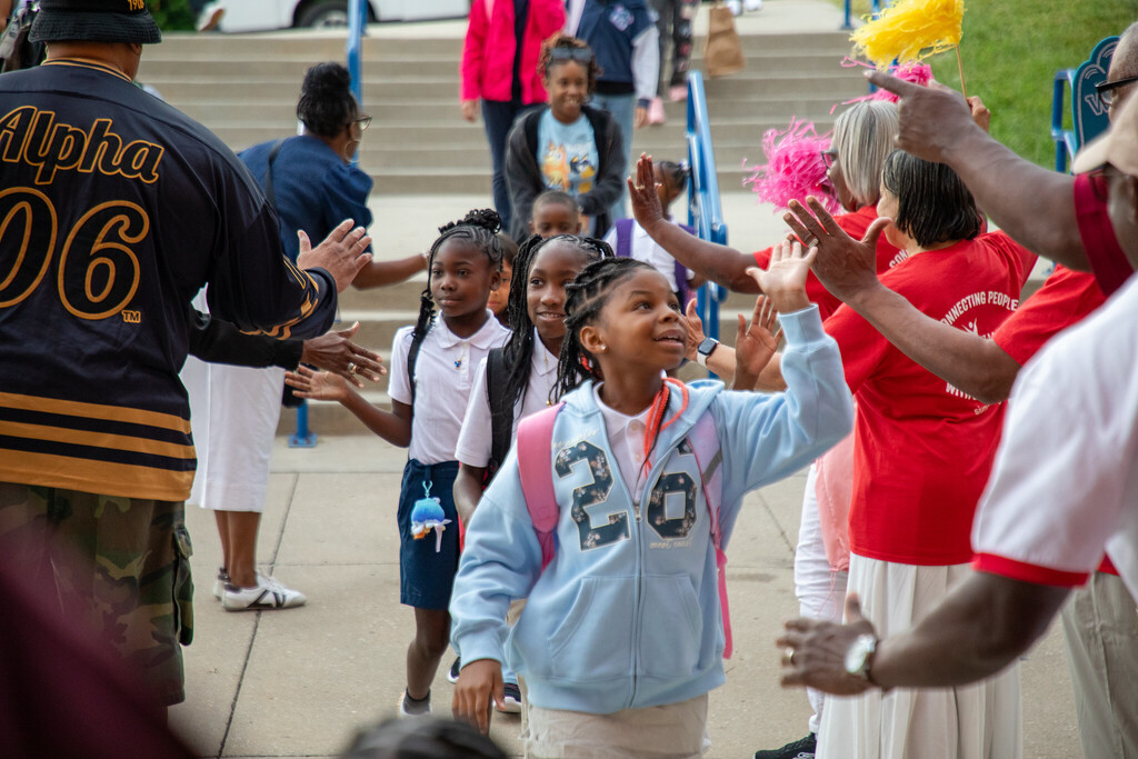 students walk toward the camera high fiving the adults who line either side of them, creating a tunnel as they walk toward a school building