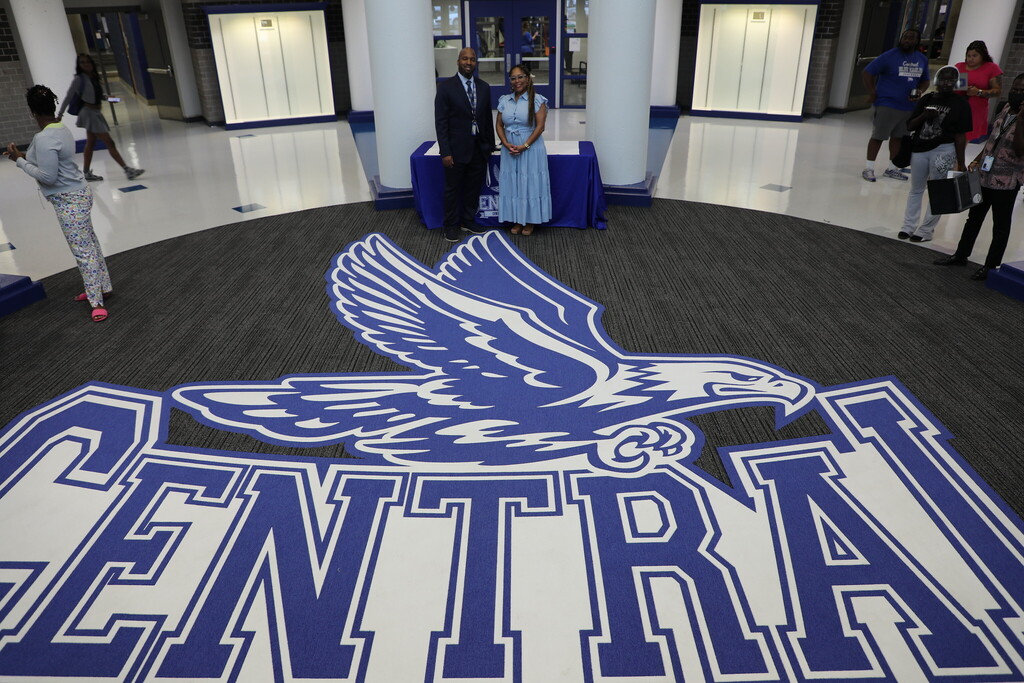 A carpet decal reading "central" with a large blue eagle on top is central in the photo, as two adults, a man and a woman, stand next to eachother smiling at the camera in the background