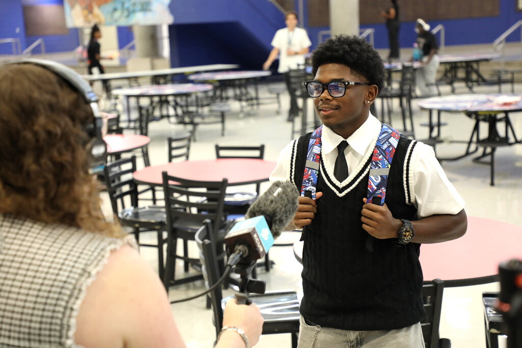 a woman faces away from the camera wearing a set of over the ear headphones and holding a microphone up to a young man who is facing the camera wearing a backpack and a black sweater vest over a white collared shirt and black tie. He wears large square glasses and a watch