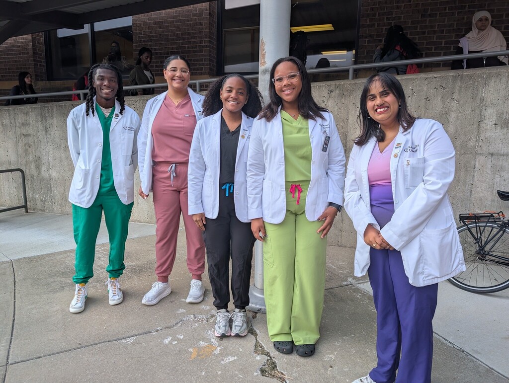four women and one young man stand next to each other wearing multicolored scrubs and white doctor coats while smiling at the camera