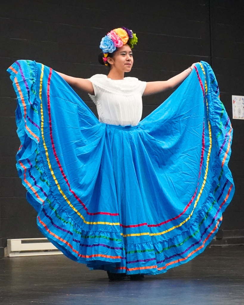 a high school girl in a traditional mexican skirt holds the skirt up at her sides with both arms and smiles at the audience