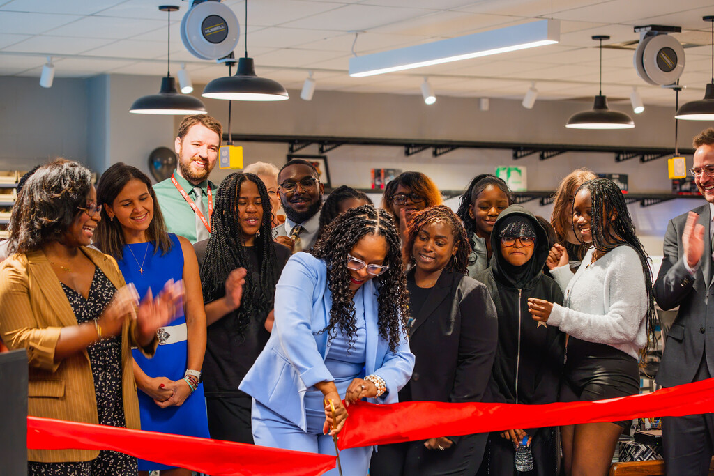 a group of people stand behind a woman in a powder blue suit as she cuts a bright red ribbon stretched in front of them