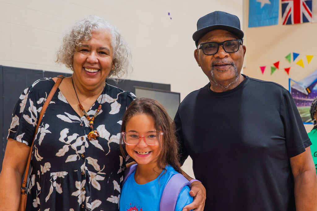 a grandma and grandpa stand on either side of their young granddaughter and all three are smiling at the camera