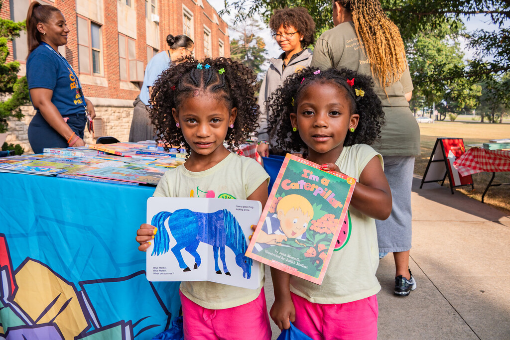 Two young girls hold children's books in front of them as they smile at the camera