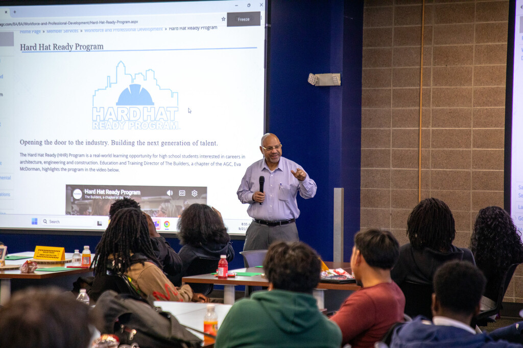 a man stands with a microphone addressing a room full of seated students
