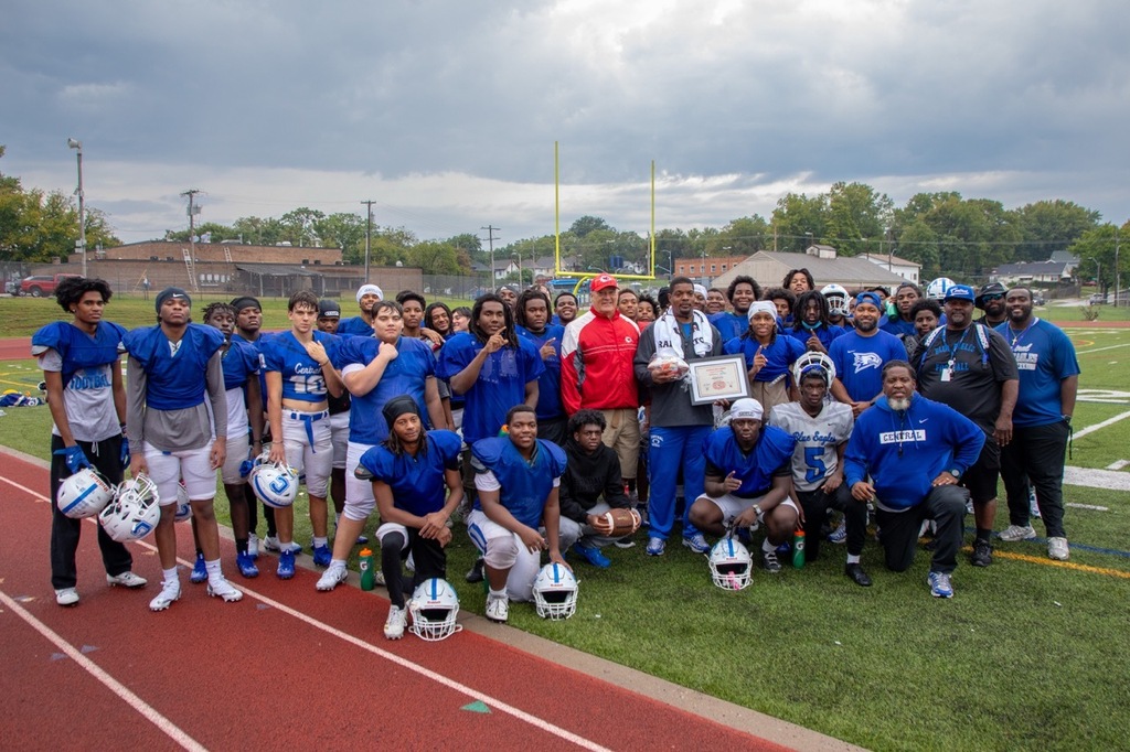 a team of football players in blue jerseys stand and kneel while holding their helmets. their coach stands in the middle holding a football and framed certificate