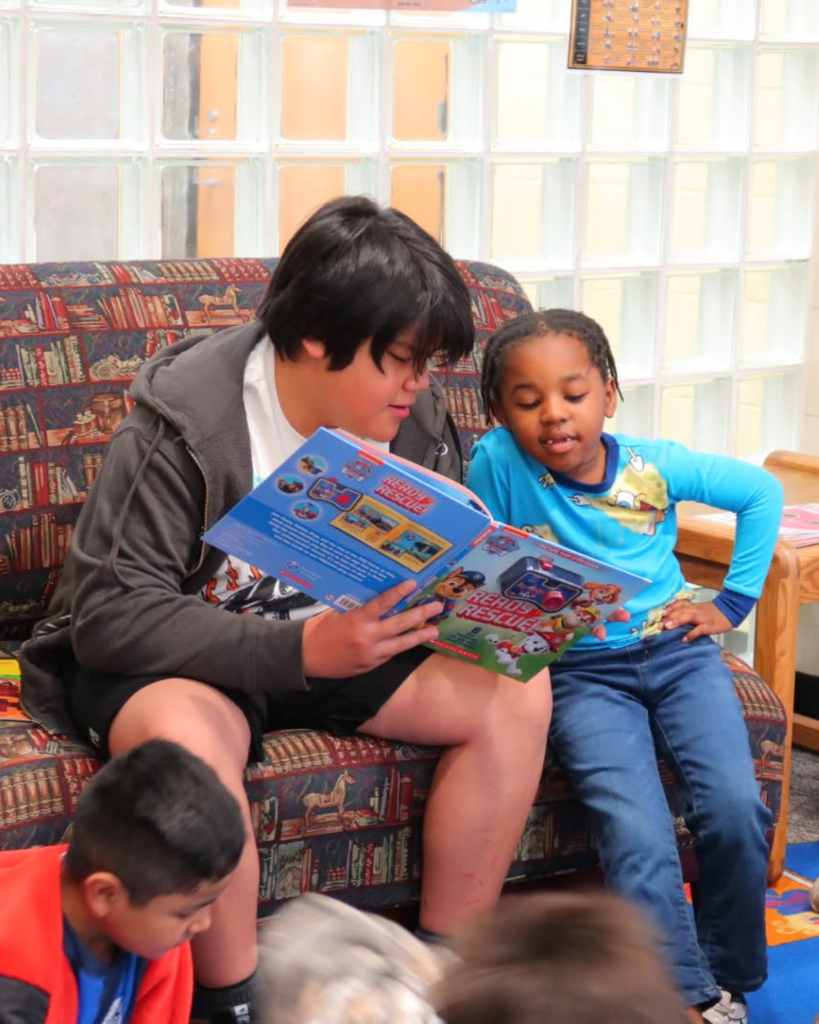 two students sit next to each other. the older student holds a book and reads to the younger student sitting next to him on a couch as other students are visible sitting on the ground in front of them doing other activities