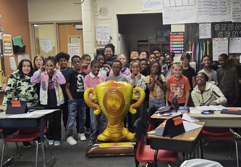 a classroom full of young students stand smiling at the camera with a large first place trophy balloon standing in front of them