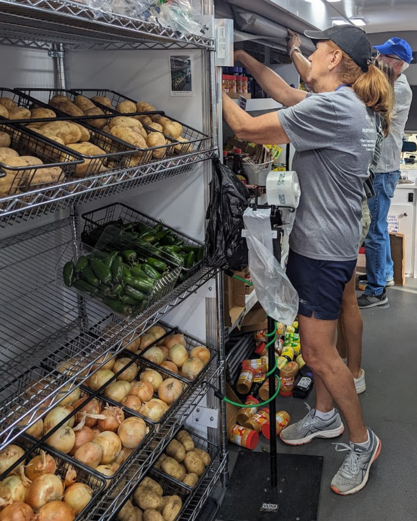 a woman in a gray t shirt, shorts and tennis shoes reaches onto a shelf holding produce and non-perishables