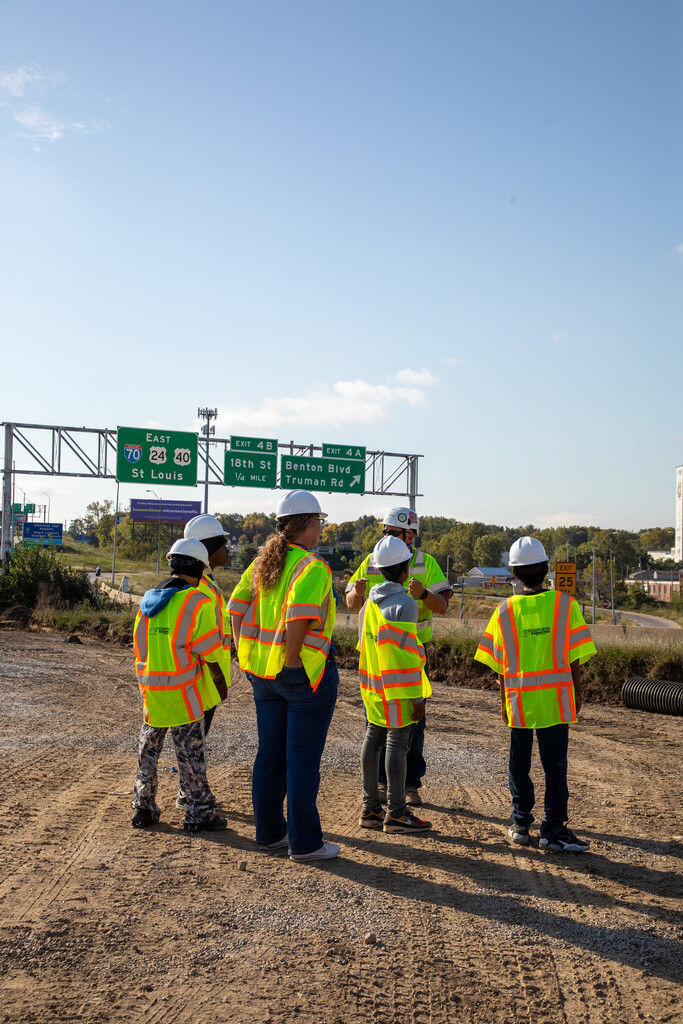 a group of young students in safety vests and construction hardhats stand on a dirt pathway with highway signs visible in the background
