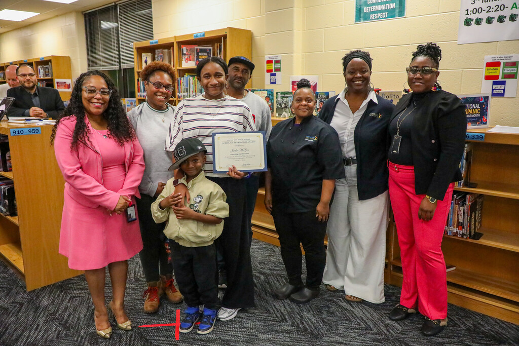 a group of adults and children stand smiling at the camera as the recipient of a superintendent award holds their certificate