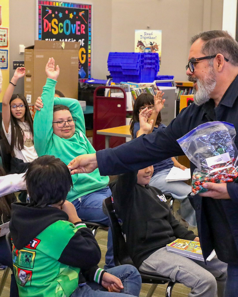 a student in a green hoodie raises his hand in the background as a man is seen handing a piece of candy to a student with her arm outstretched