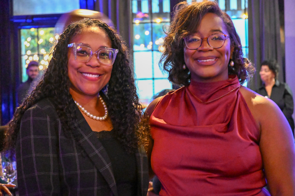 two women stand next to each other smiling at the camera. they each wear glasses, the woman on the left has long hair and is wearing a black blazer with a plaid design and a pearl necklace, the woman on the right wears a red cowl-neck top and has shoulder length hair