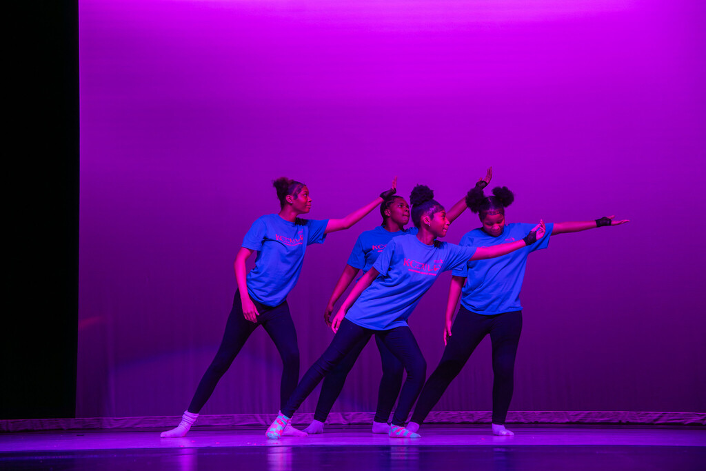 a group of four female students wearing matching blue t-shirts and dark colored pants dance on a stage in unison