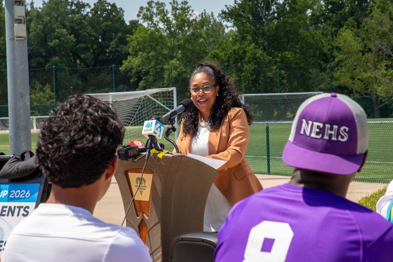 Superintendent Dr. Jennifer collier stands at a podium to address a crowd and is looking toward two male KCPS student-athletes sitting in chairs. One wears a purple "NEHS" baseball hat backwards with the type visible to the camera