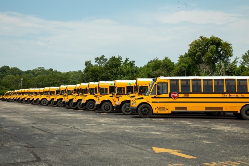 a long line of school buses stretches into the distance. they are all parked next to each other, and have 'zum services, inc' written on the side. green trees and a moderately cloudy sky are visible behind them