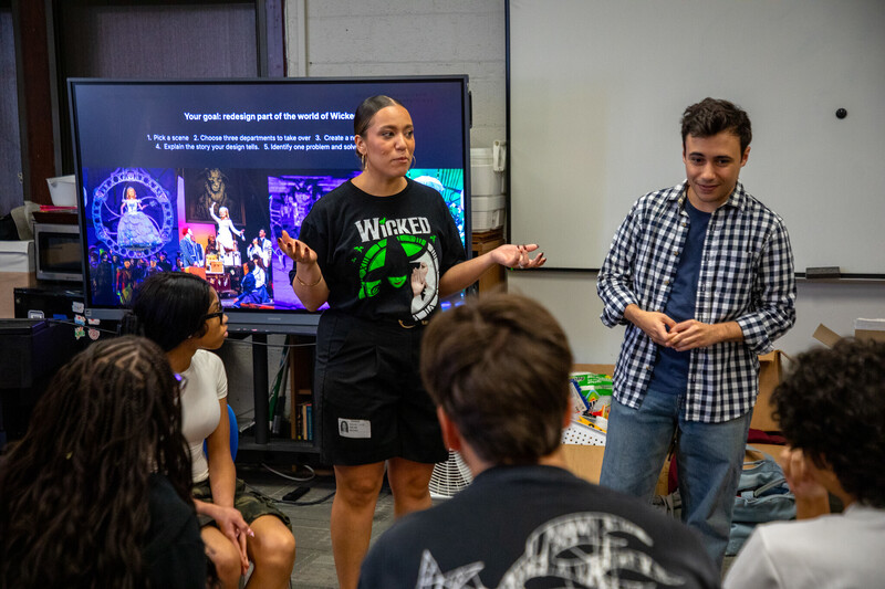 a woman wearing a black t shirt with the logo for the musical wicked on the front speaks to a group of teenagers who are facing toward her and away from the camera. next to her, a man in a checkered button up shirt smiles at the teens