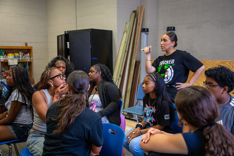 a group of teens sitting in chairs look up listening to an adult woman who is speaking with one hand on her hip and the other hand gesticulating in the air. she wears a black t shirt with the wicked musical logo on the front.