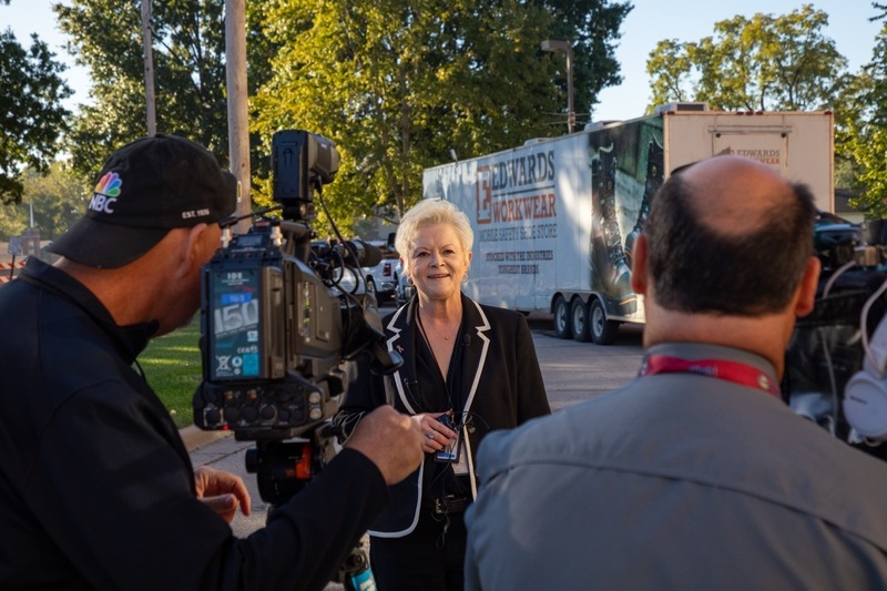 a woman in a black blazer with white trim stands in front of two television cameras giving an interview