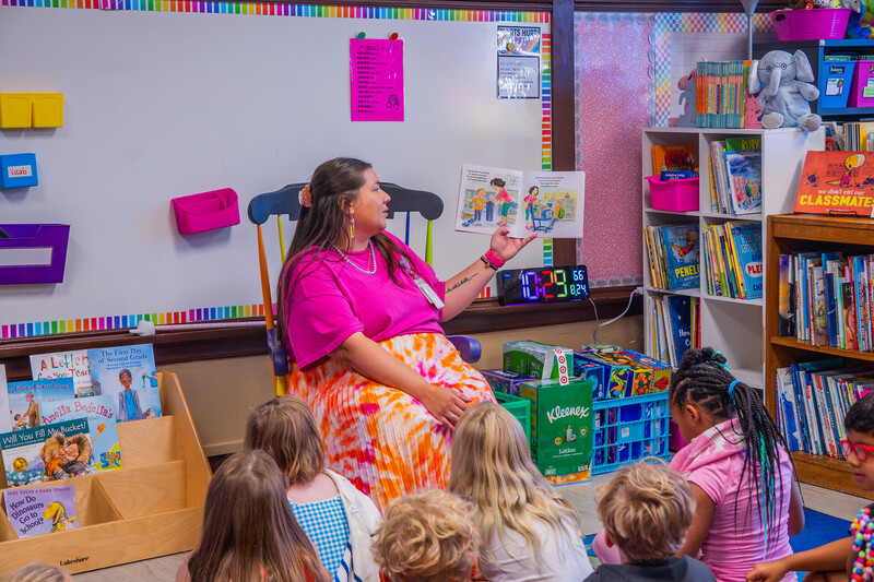 a woman in a pink top and multicolored skirt sits in a rocking chair reading a picture book to a group of students seated on the floor in front of her