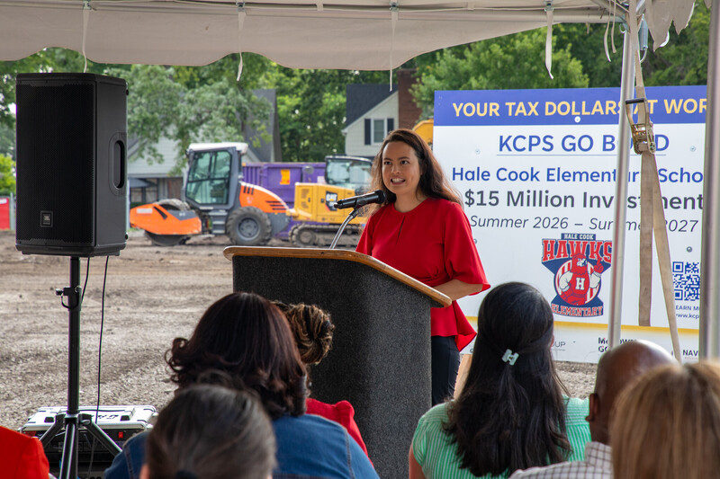 a woman in a red top stands at a podium addressing a crowd as dirt and construction equipment is visible in the background