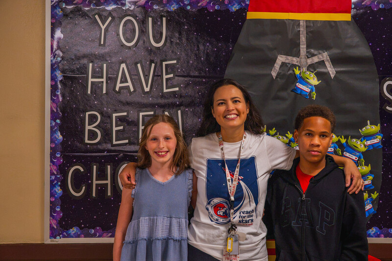 a woman stands smiling with her arms around two students on either side of her, one smiles and the other looks at the camera