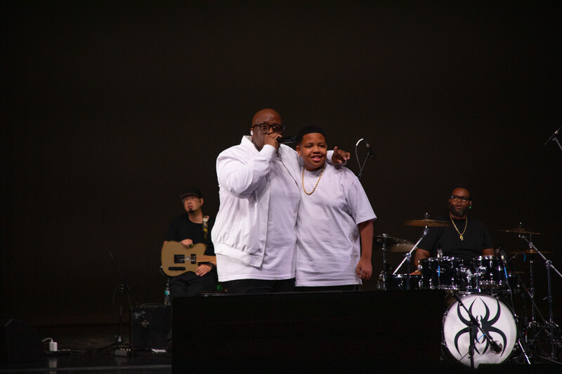 a man in a white t shirt and white jacket holds a microphone in one hand and has his other arm around a boy, his son, who is wearing a white t shirt and gold chain