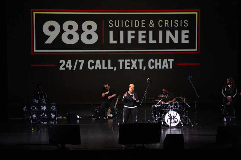 a band and woman stand on a stage with information for the 988 suicide and crisis lifeline projected onto a large screen behind them