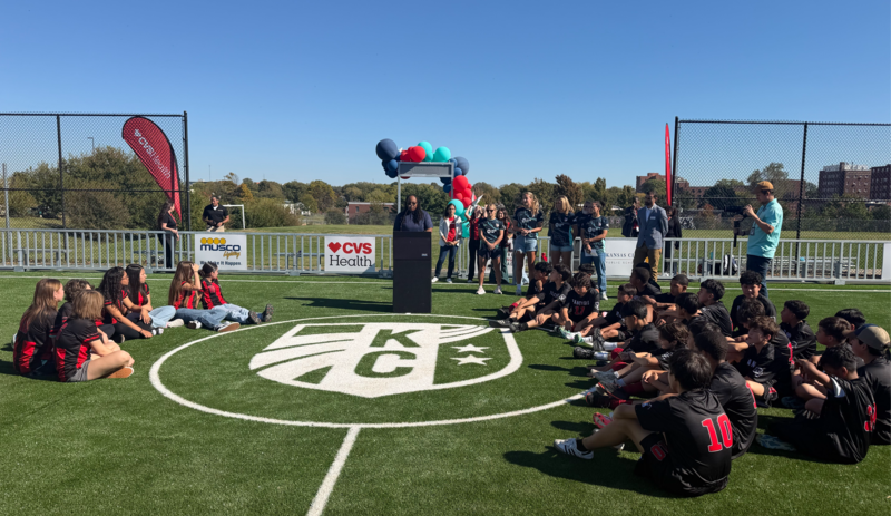several students sit on a turf soccer field looking toward a podium as a speaker addresses the crowd