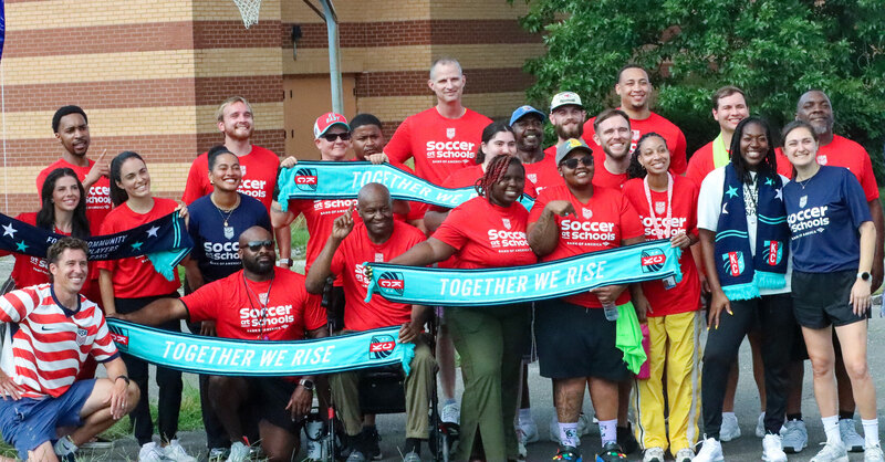 adults wearing red soccer in schools branded t-shirts stand smiling at the camera together. some hold teal colored scarves that say together we rise