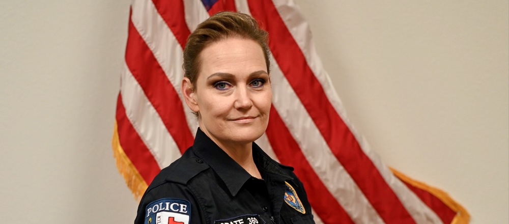woman sitting in a three quarter pose in her officer uniform smiling with an American flag behind her