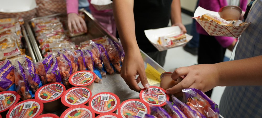 photo of hands of students grabbing applesauce and holding small food plates 