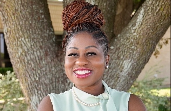 dark skinned woman with arms crossed smiling at the camera standing in front of a tree