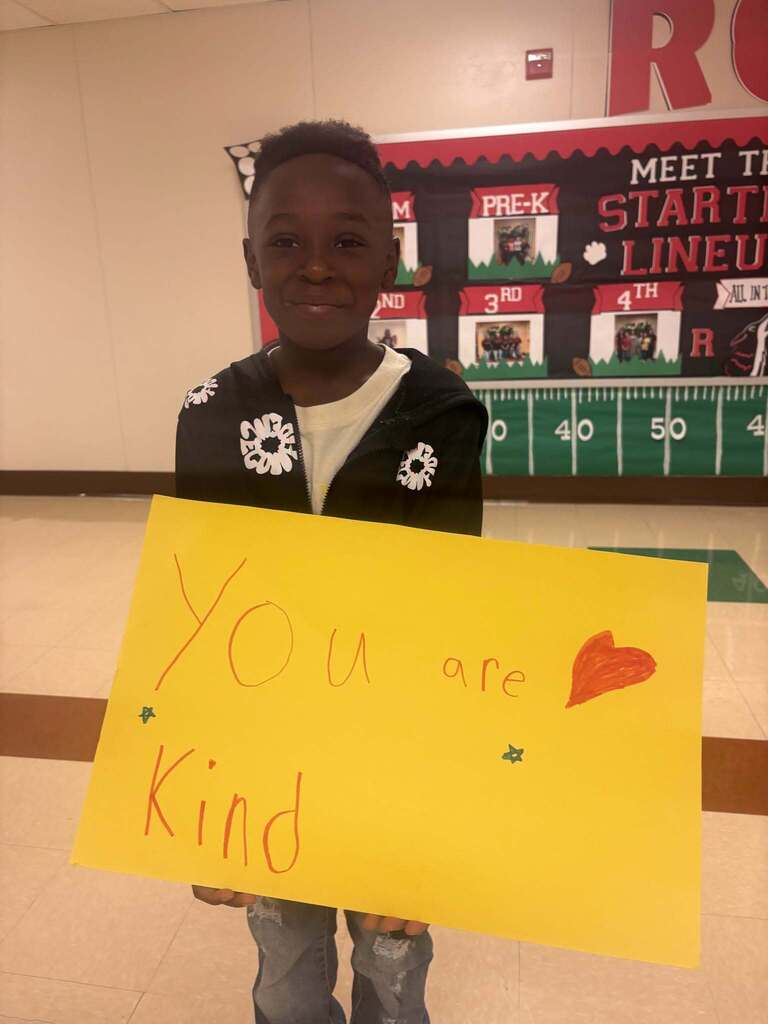 A child holds a yellow sign with "YOU are kind" written on it. A football field banner is in the background.