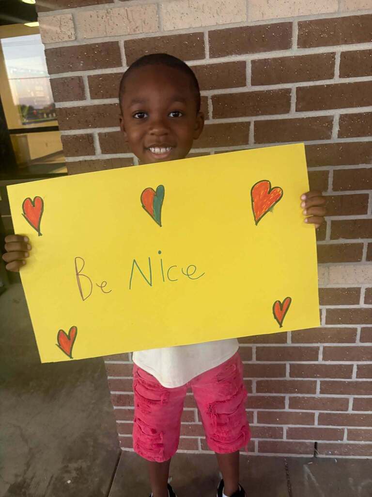 A smiling child holds a yellow sign with red hearts and the message "Be Nice" against a brick wall.