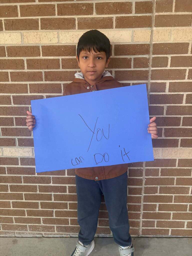 A child holds a blue sign with white writing, "You can do it," in front of a brick wall.